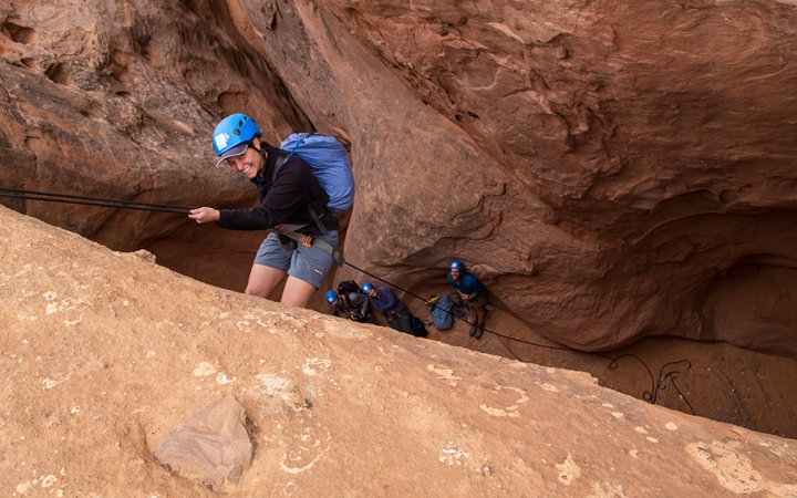 A person wearing safety gear is secured by ropes as they descend into a canyon. They are looking up at the camera and smiling.